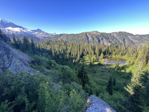 A scenic view of a lush green valley with a small pond, surrounded by mountains and trees under a clear blue sky.