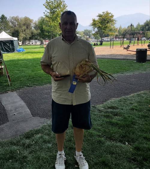 A man stands in a park holding a large vegetable and a trophy, with trees and playground equipment in the background.