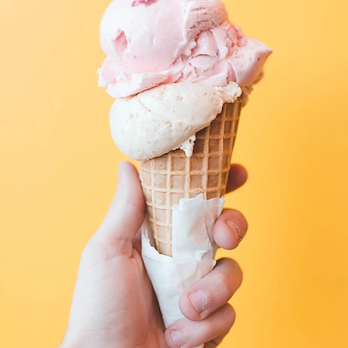 A hand holding a waffle cone with two scoops of pink and white ice cream against a bright yellow background.