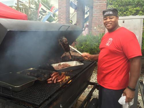 A smiling man in a red shirt grills meat on a barbecue, surrounded by smoke and outdoor decorations.