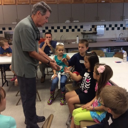 A man demonstrates something to a group of children sitting at a table in a classroom setting.
