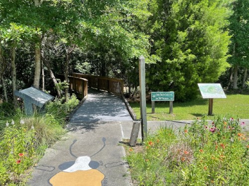 A wooden bridge leads into a lush, green area with informational signs and colorful flowers along the path.