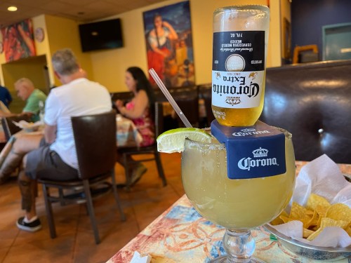 A margarita glass topped with a Corona beer, with people dining in the background. Chips are on the table.