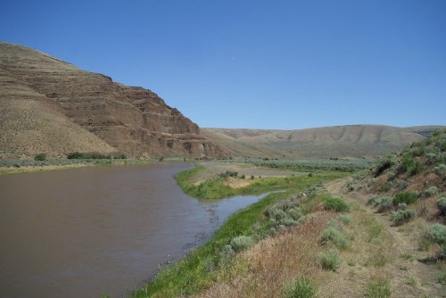 A serene river flows alongside grassy banks and rocky hills under a clear blue sky.