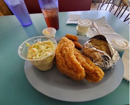 A plate with fried fish, coleslaw, a baked potato in foil, and two dipping sauces, with drinks in the background.