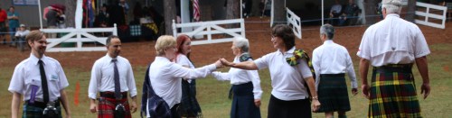 A group of people in kilts engaging in a friendly handshake at an outdoor event. Flags and tents are in the background.