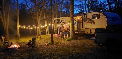A cozy camper surrounded by trees at dusk, with string lights and a campfire nearby, creating a warm atmosphere.