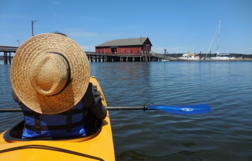 A person in a straw hat paddles a kayak on calm water, with a red building and boats in the background.