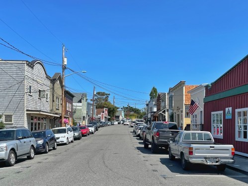 A sunny street lined with parked cars and quaint shops, under a clear blue sky.