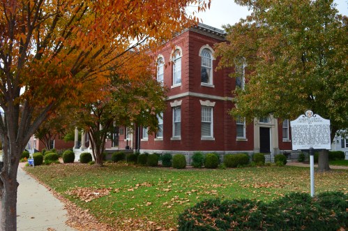 A brick building surrounded by colorful autumn trees and fallen leaves, with a historical marker in the foreground.