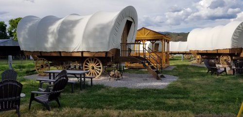 Two covered wagons on a grassy area, with wooden steps and seating nearby, set against a cloudy sky.