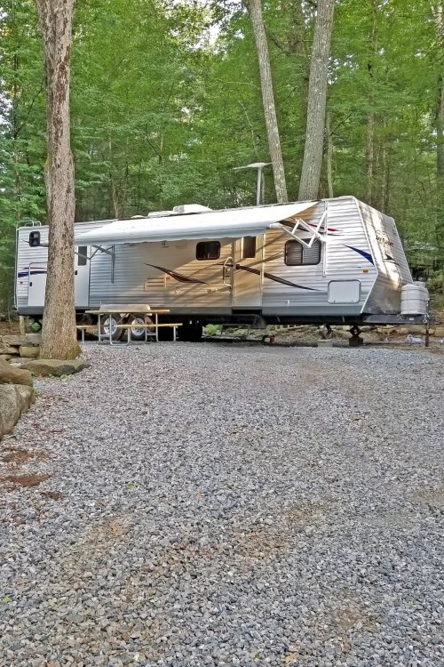 A silver travel trailer parked on a gravel site surrounded by trees in a wooded area.