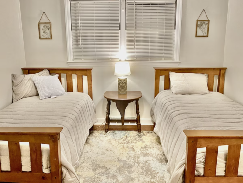 A cozy bedroom with two wooden beds, a small table, and a lamp, featuring light-colored bedding and soft decor.