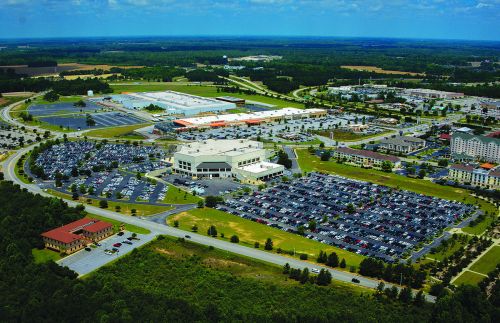 Aerial view of a large commercial area with parking lots, buildings, and green spaces under a blue sky.