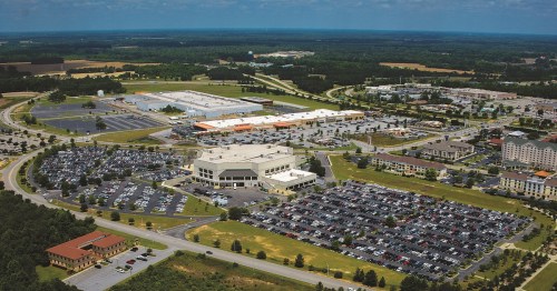 Aerial view of a large commercial area with multiple buildings, parking lots, and green spaces under a blue sky.