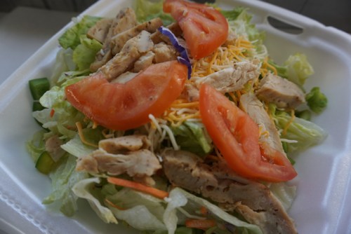 A fresh salad topped with grilled chicken, tomatoes, shredded cheese, and mixed greens in a takeout container.