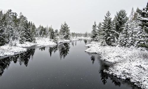 A serene winter landscape featuring a snow-covered river surrounded by trees, reflecting the snowy scenery.