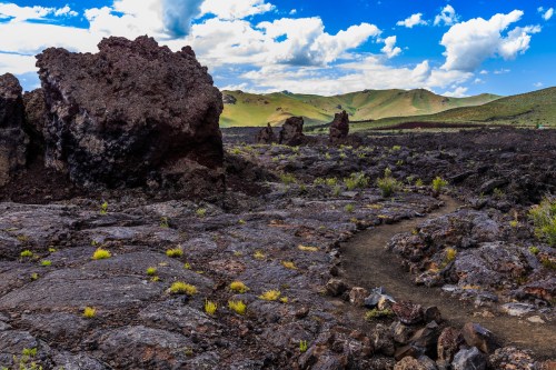 A rocky volcanic landscape with large boulders, a winding path, and green hills under a blue sky with clouds.