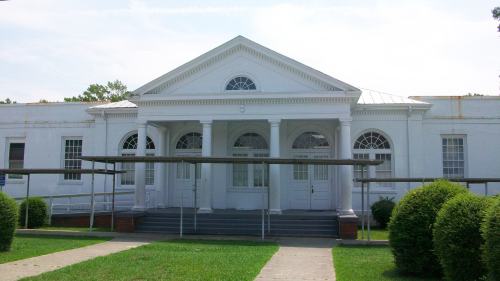 A white building with a pediment and columns, featuring large windows and a grassy area in front.
