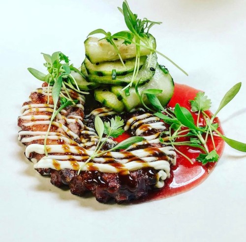 A beautifully plated dish featuring octopus, cucumber, and garnishes, drizzled with sauces on a white background.