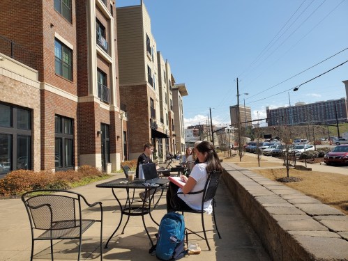 Two people work on laptops at outdoor tables near an apartment building on a sunny day.