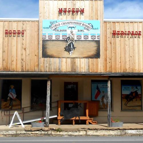 Front view of a wooden museum building with rodeo-themed murals and signage, featuring a bench outside.