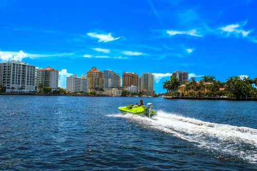 A person rides a bright green jet ski on a calm waterway, with city buildings and palm trees in the background under a blue sky.