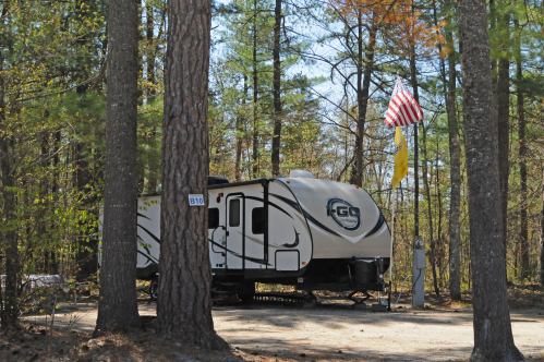 A camper parked in a wooded area, with an American flag and a yellow flag nearby, surrounded by tall trees.