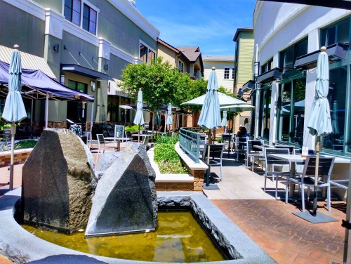 A sunny outdoor dining area with a small fountain, tables, and umbrellas surrounded by greenery and buildings.