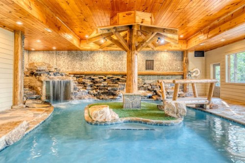 Indoor pool area featuring a wooden ceiling, stone waterfall, and a central wooden structure surrounded by water.
