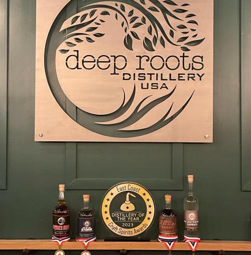 Sign for Deep Roots Distillery with award plaque and four bottles of spirits displayed in front.
