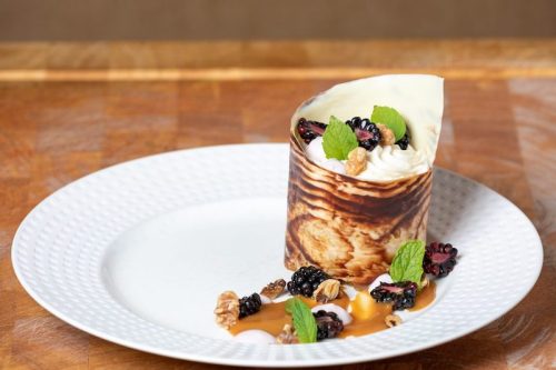 A dessert in a rolled chocolate cup, topped with cream, blackberries, nuts, and mint, served on a white plate.