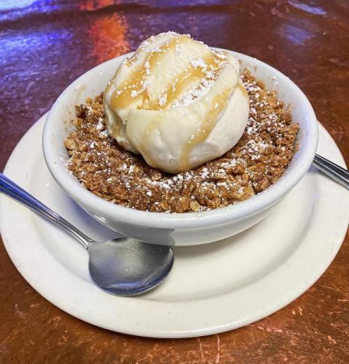 A bowl of warm dessert topped with vanilla ice cream and drizzled with caramel sauce, served with a spoon.