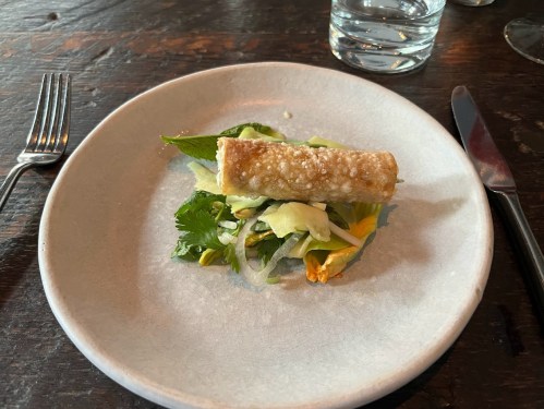 A plate with a crispy roll on a bed of fresh greens and vegetables, set on a rustic wooden table.