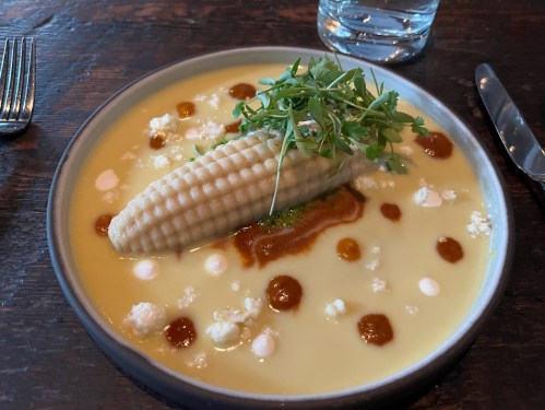 A bowl of creamy corn soup topped with a corn cob, herbs, and dollops of sauce, served on a wooden table.