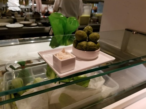 A small plate with green spherical desserts and a side of creamy sauce, displayed on a glass shelf in a restaurant.