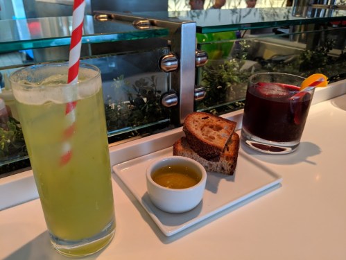 A refreshing green drink with a straw, a glass of dark beverage, and a plate of bread with olive oil on a white table.