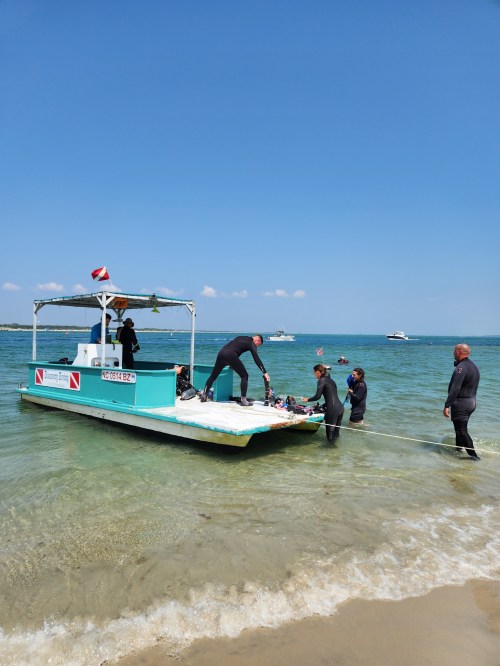 A dive boat anchored near the shore with divers preparing equipment in clear blue water under a sunny sky.