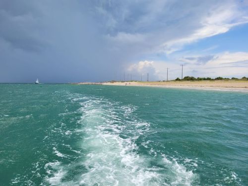 A view of a turquoise sea with a boat's wake, dark clouds looming over a sandy beach in the distance.