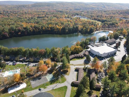 Aerial view of a serene lake surrounded by autumn foliage and a resort with buildings and a pool nearby.
