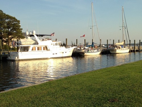 A white yacht and several sailboats docked along a calm waterway, with green grass and trees in the background.