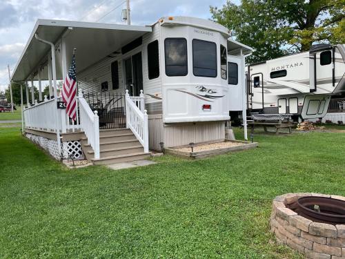 A parked RV with a porch, steps, and an American flag, surrounded by grass and another RV in the background.