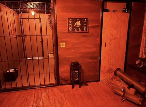 A dimly lit room with a barred window, wooden walls, and a door, featuring a small table and a cannon.