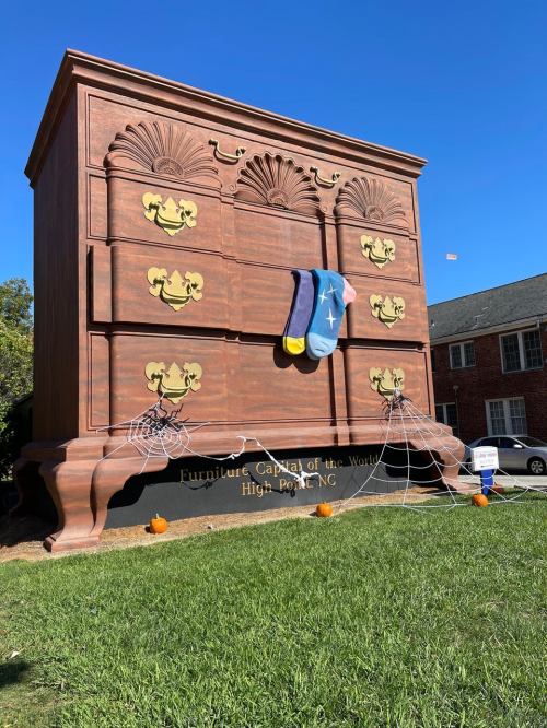 A giant wooden dresser with colorful socks hangs from the front, set against a clear blue sky in High Point, NC.