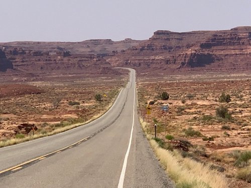 A long, straight road stretches through a desert landscape with red rock formations in the background.