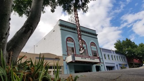 Historic California theater with a colorful facade and marquee, surrounded by greenery and under a partly cloudy sky.