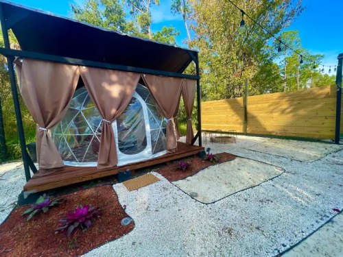 A cozy outdoor geodesic dome with curtains, surrounded by gravel and plants, set in a natural landscape.