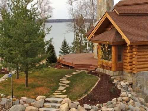 A cozy log cabin by a lake, surrounded by trees and a stone pathway leading to a wooden deck.