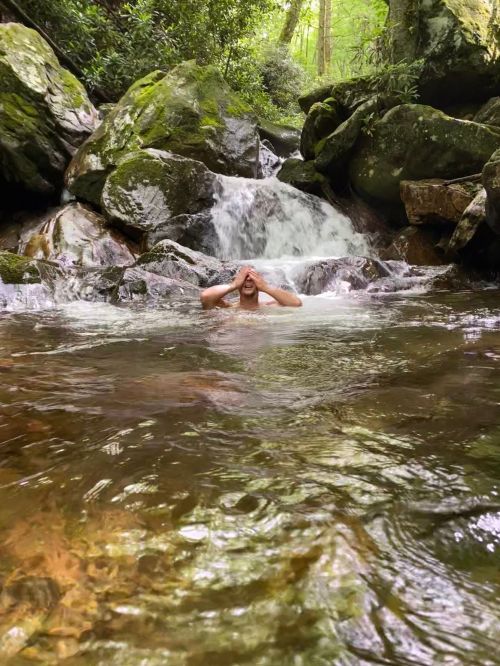 A person is submerged in a serene stream, surrounded by rocks and lush greenery, with a small waterfall in the background.