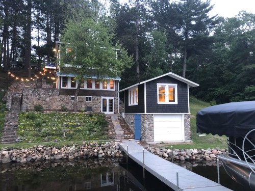 A charming lakeside cabin with stone accents, surrounded by trees and a dock leading to the water.